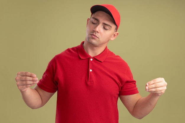 Foto gratuita colpito il giovane fattorino che indossa l'uniforme e il berretto che finge di tenere qualcosa di isolato sulla parete verde oliva