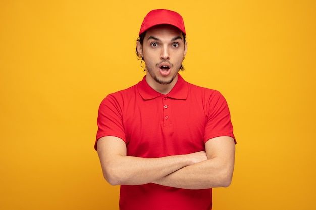 impressed young delivery man wearing uniform and cap standing with crossed arms looking at camera isolated on yellow background