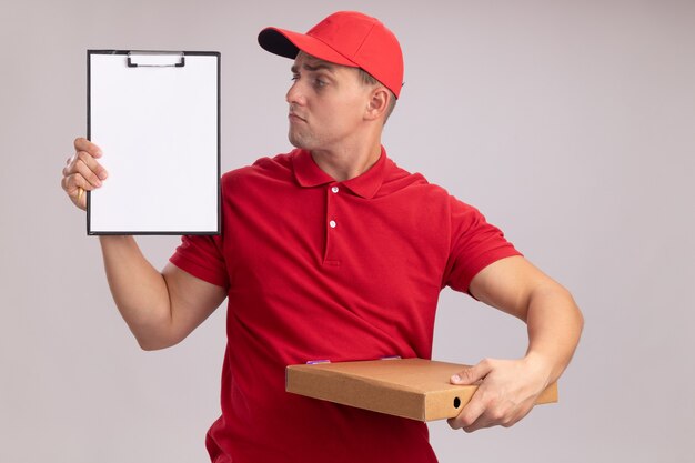 Впечатленный молодой курьер в униформе с кепкой, держащей коробку для пиццы и смотрящий в буфер обмена в руке, изолированной на белой стене