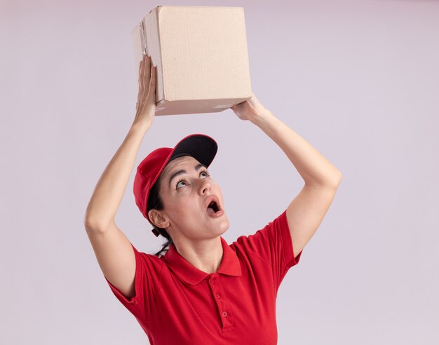 Free photo impressed young delivery woman in uniform and cap holding cardbox above head looking at it