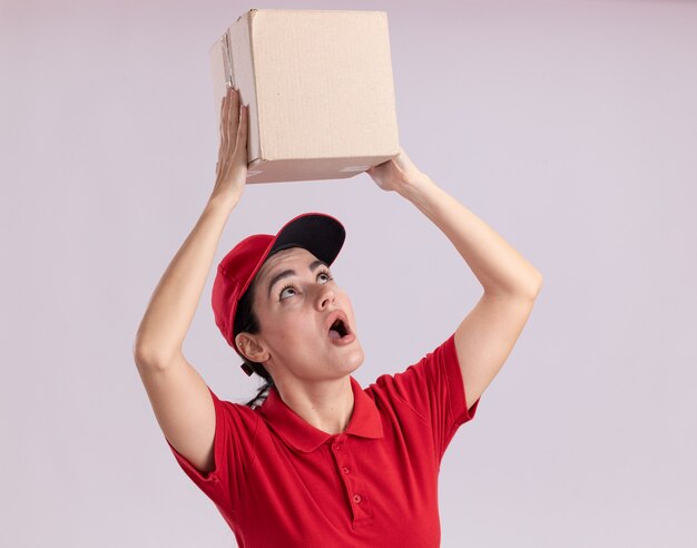Impressed young delivery woman in uniform and cap holding cardbox above head looking at it 