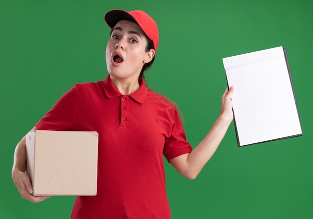 Impressed young delivery woman in uniform and cap holding cardbox and showing clipboard looking at front isolated on green wall