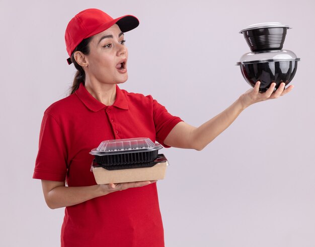 Impressed young delivery woman in uniform and cap holding paper food package and food containers looking at food containers 