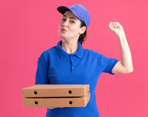 Impressed young delivery woman in uniform and cap holding pizza packages  doing knocking gesture isolated on pink wall