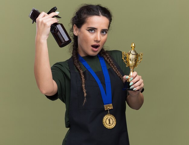 Впечатленная молодая женщина-парикмахер в униформе и медали, держащая кубок победителя и поднимающая инструменты парикмахера, изолированные на оливково-зеленой стене