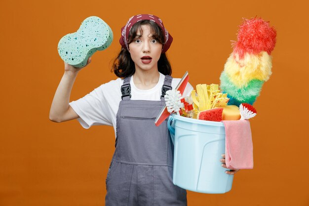 Impressed young female cleaner wearing uniform and bandana holding bucket of cleaning tools looking at camera rising sponge up isolated on orange background