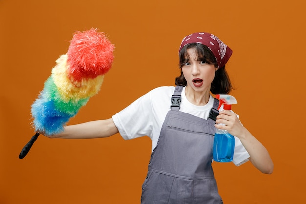 Впечатленная молодая женщина-уборщица в униформе и бандане, держащая моющее средство, смотрящая в камеру, растягивая тряпку из перьев на оранжевом фоне