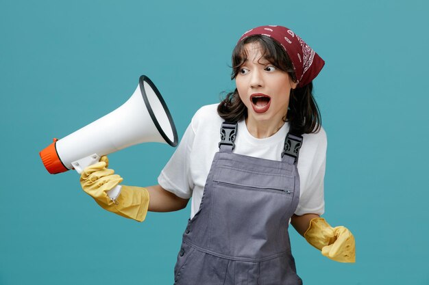 制服のバンダナとゴム手袋を身に着けている印象的な若い女性クリーナーは、青い背景で隔離された側を見て自分自身にそれを指しているスピーカーを保持しています