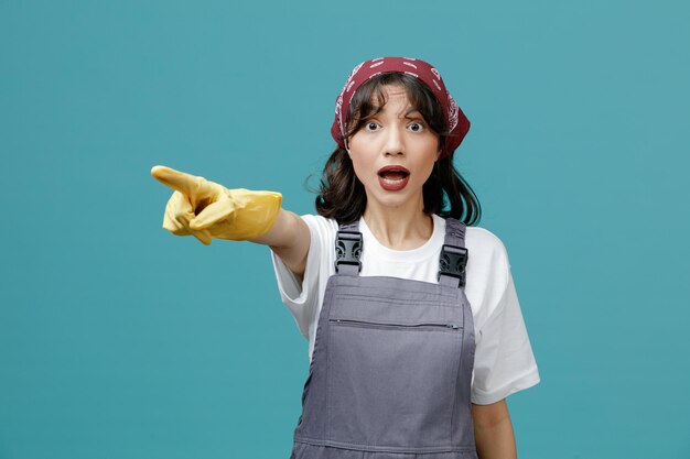 青い背景で隔離の側を指しているカメラを見て制服バンダナとゴム手袋を身に着けている印象的な若い女性クリーナー