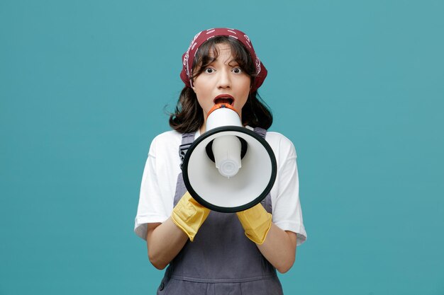 impressed young female cleaner wearing uniform bandana and rubber gloves looking at camera talking into speaker isolated on blue background