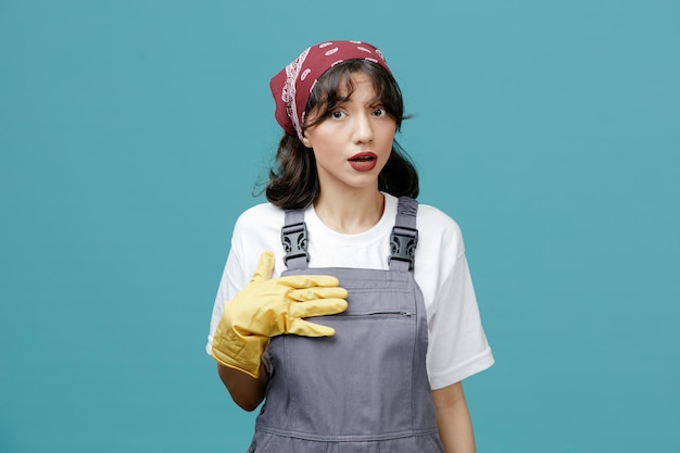 Impressionata giovane donna addetta alle pulizie che indossa bandana uniforme e guanti di gomma che puntano su se stessa con la mano che guarda la fotocamera isolata su sfondo blu