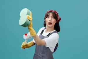 Free photo impressed young female cleaner wearing uniform bandana and rubber gloves standing in profile view holding toilet brushes stretching sponge out looking at camera pretend cleaning