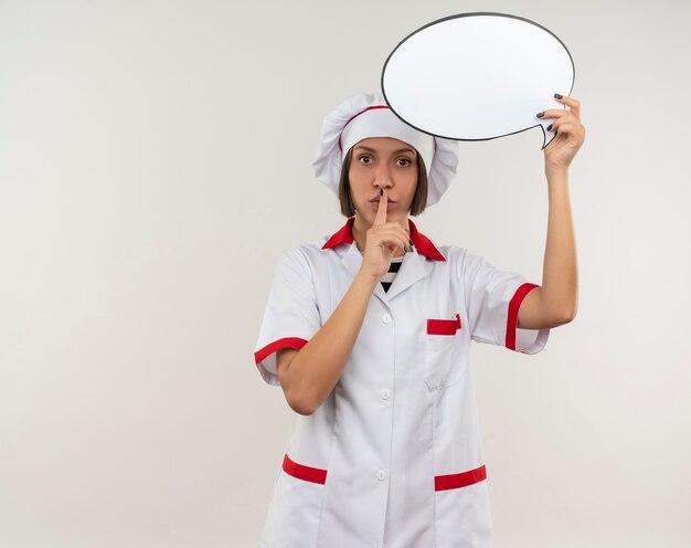 Impressed young female cook in chef uniform holding chat bubble and gesturing silence isolated on white  with copy space