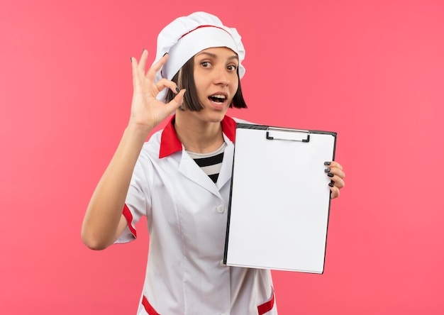 Impressed young female cook in chef uniform holding clipboard and doing ok sign isolated on pink 