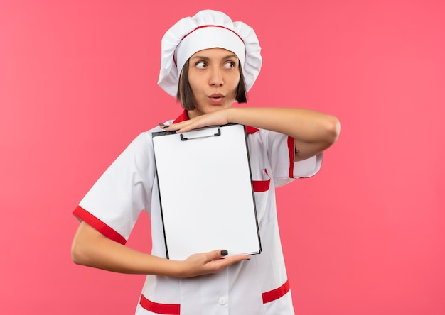 Foto gratuita impressionato giovane cuoco femminile in uniforme del cuoco unico che tiene appunti e guardando il lato isolato sul rosa