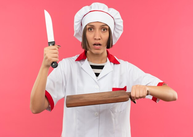 Впечатленная молодая женщина-повар в униформе шеф-повара держит нож и разделочную доску, изолированные на розовом