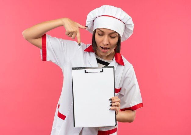 Free photo impressed young female cook in chef uniform holding looking and pointing at clipboard isolated on pink  with copy space