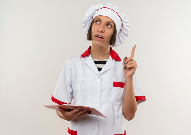 메모 패드를 들고 복사 공간이 흰색에 고립 된 측면을보고 손가락을 올리는 요리사 유니폼에 감동 된 젊은 여성 요리사