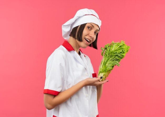 Impressed young female cook in chef uniform holding and pointing with hand lettuce isolated on pink 