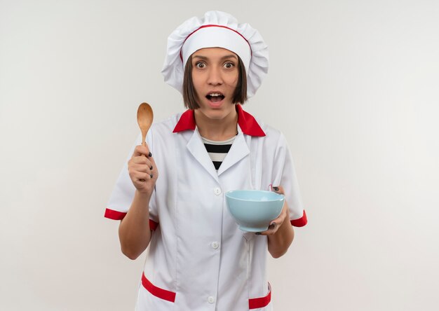 Impressed young female cook in chef uniform holding spoon and bowl isolated on white  with copy space