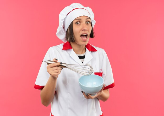 Впечатленная молодая женщина-повар в униформе шеф-повара держит венчик и миску, изолированные на розовом с копией пространства