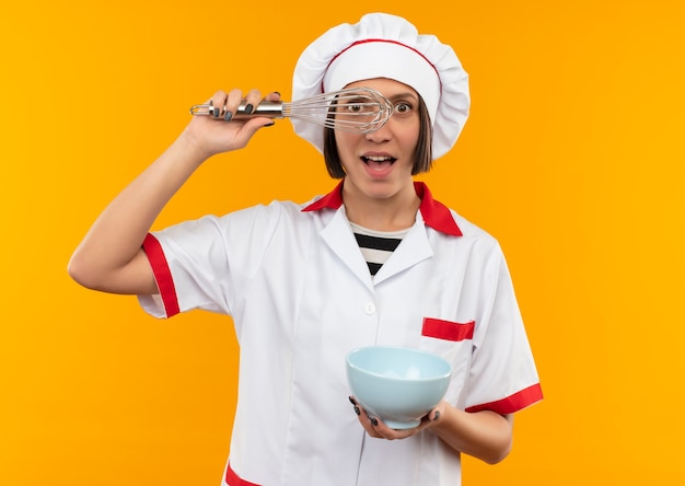 Impressed young female cook in chef uniform holding whisk and bowl looking through whisk isolated on orange 