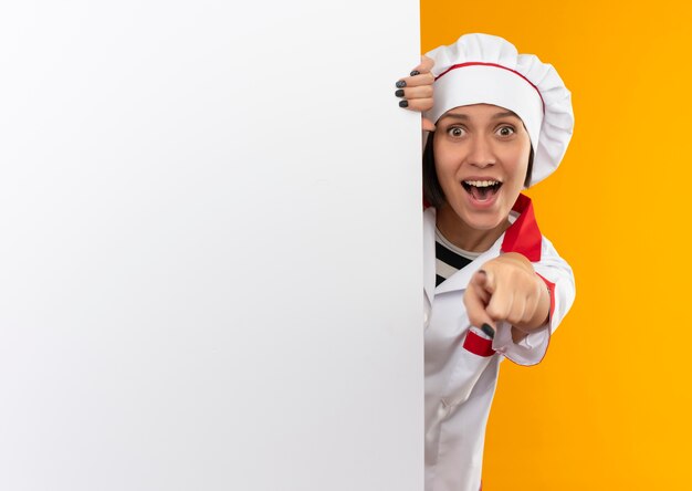 Впечатленная молодая женщина-повар в униформе шеф-повара, стоящая за белой стеной и указывая на оранжевый с копией пространства