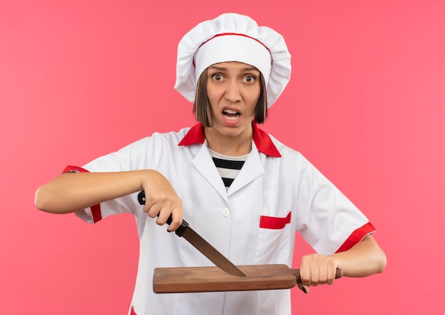 Впечатленная молодая женщина-повар в униформе шеф-повара трогает разделочную доску с ножом, изолированным на розовом
