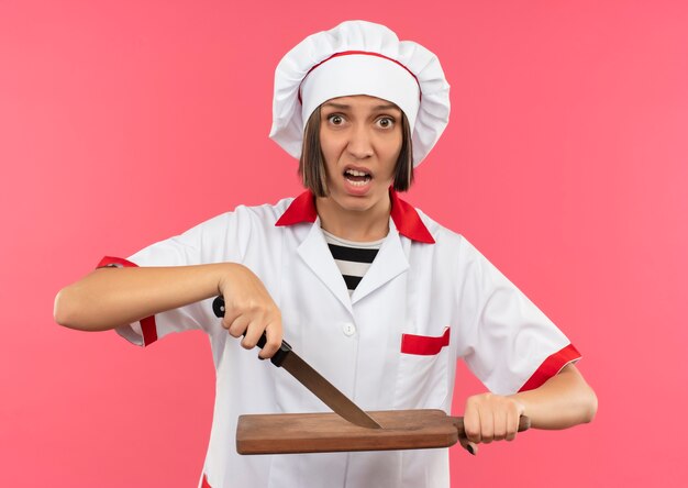 Impressed young female cook in chef uniform touching cutting board with knife isolated on pink 