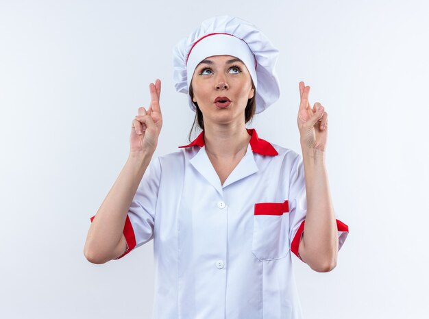 Impressed young female cook wearing chef uniform crossing fingers isolated on white wall