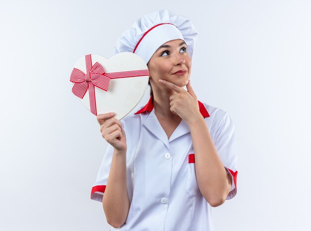 impressed young female cook wearing chef uniform holding heart shape box grabbed chin isolated on white wall