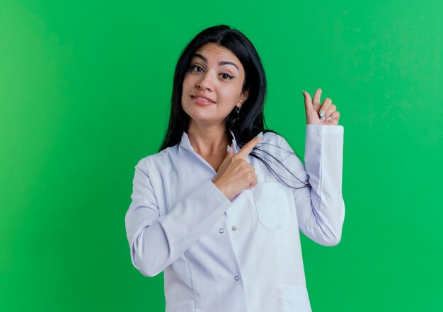 Impressed young female doctor wearing medical robe looking pointing behind 