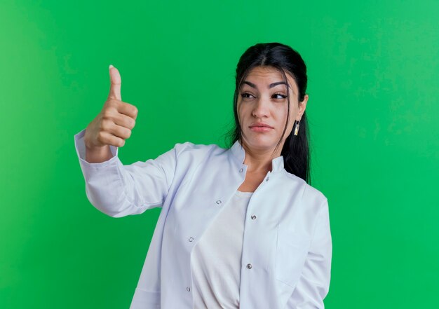 Impressed young female doctor wearing medical robe looking at side showing thumb up isolated on green wall with copy space