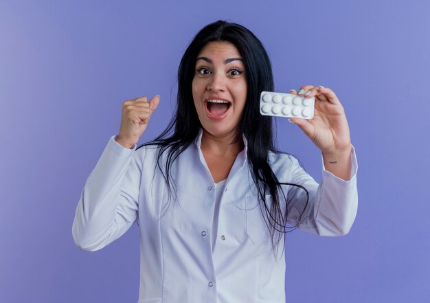 Impressed young female doctor wearing medical robe showing pack of medical tablets, looking clenching fist 