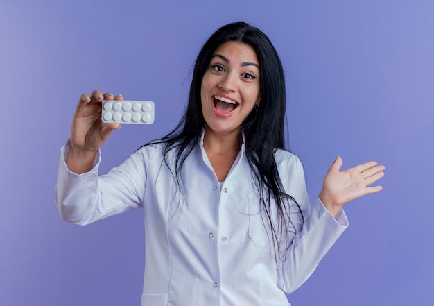 Впечатленная молодая женщина-врач в медицинском халате, показывающая упаковку медицинских таблеток, глядя, показывая пустую руку