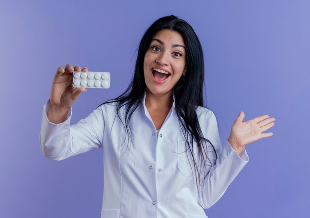 Impressed young female doctor wearing medical robe showing pack of medical tablets, looking showing empty hand 