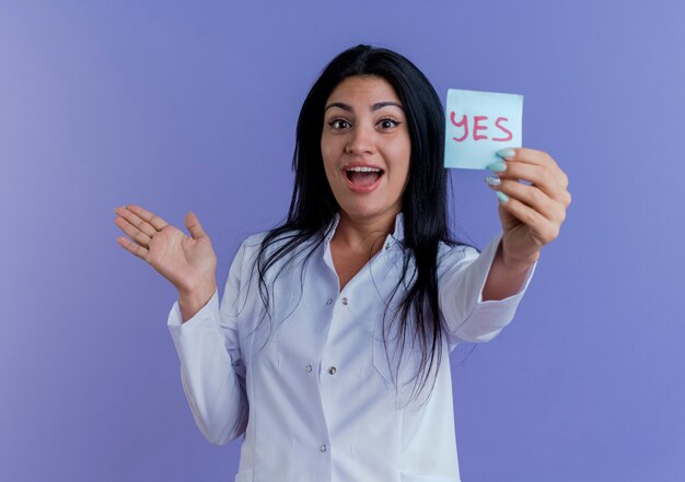 Impressed young female doctor wearing medical robe showing yes note showing empty hand isolated on purple wall with copy space