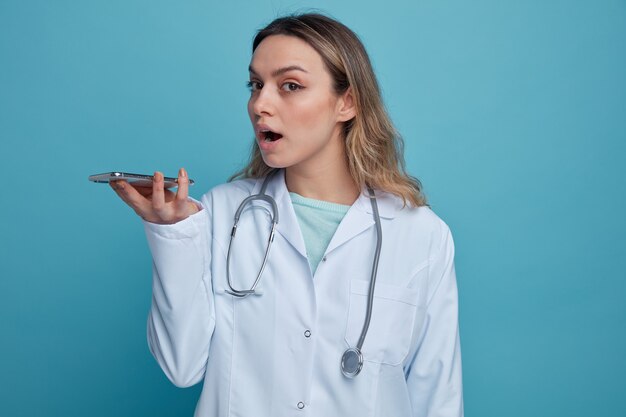 Impressed young female doctor wearing medical robe and stethoscope around neck holding mobile phone horizontally talking by its microphone 