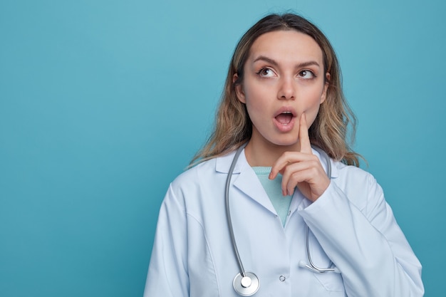 Impressed young female doctor wearing medical robe and stethoscope around neck touching cheek with finger looking up 