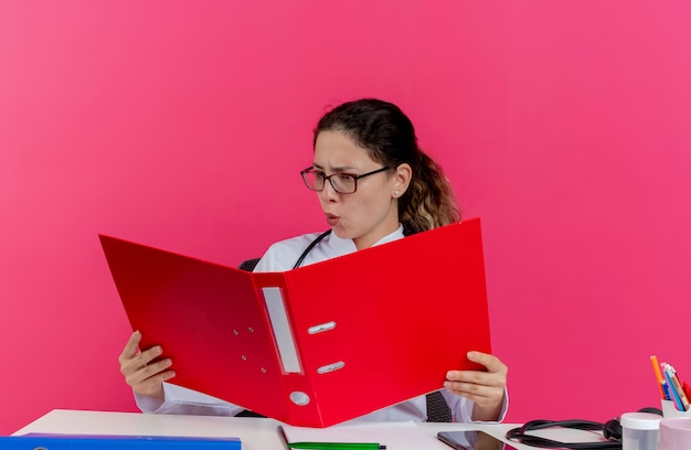 Impressionato giovane medico femminile che indossa abito medico e stetoscopio e occhiali seduto alla scrivania con strumenti medici che tengono e guardando la cartella isolata