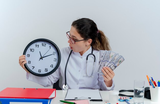 impressed young female doctor wearing medical robe and stethoscope and glasses sitting at desk with medical tools holding money and clock and looking at clock isolated