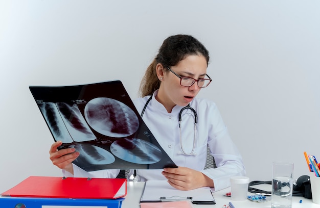 Impressed young female doctor wearing medical robe and stethoscope and glasses sitting at desk with medical tools holding x-ray shot looking down isolated