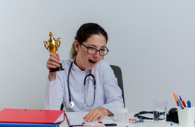 Free photo impressed young female doctor wearing medical robe and stethoscope and glasses sitting at desk with medical tools looking down holding winner cup putting hand on desk isolated