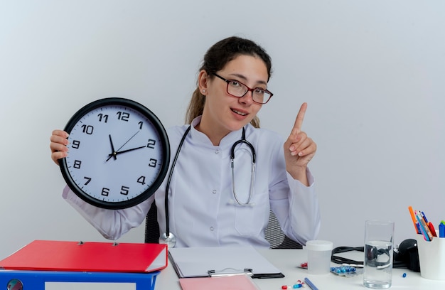 Impressed young female doctor wearing medical robe and stethoscope and glasses sitting at desk with medical tools looking holding clock raising finger isolated
