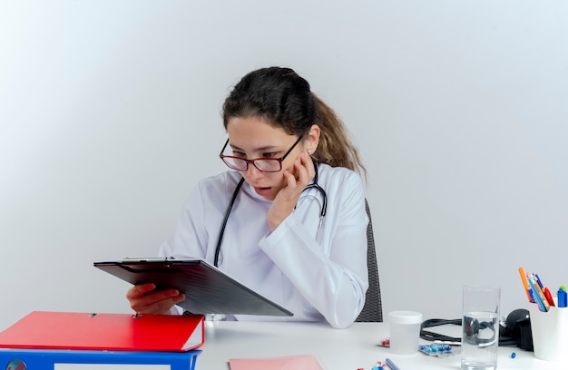 Foto gratuita impressionato giovane medico femminile che indossa veste medica e stetoscopio e occhiali seduto alla scrivania con strumenti medici toccando il viso tenendo e guardando negli appunti isolati