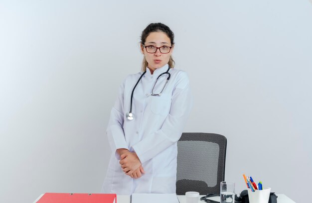 Impressed young female doctor wearing medical robe and stethoscope and glasses standing behind desk with medical tools looking keeping hands together isolated