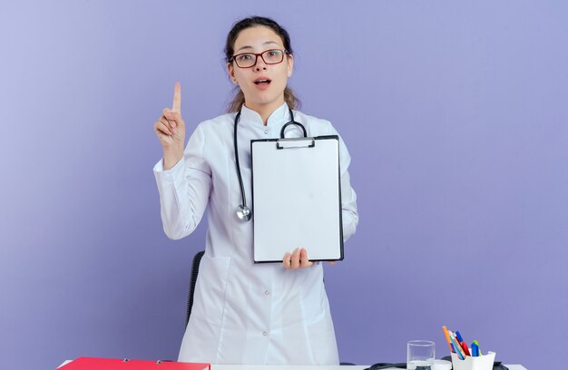 의료 가운 및 청진 기 및 의료 도구를 책상 뒤에 서있는 안경을 착용하는 감동 된 젊은 여성 의사 절연 클립 보드를 보여주는 제기 손가락을 찾고