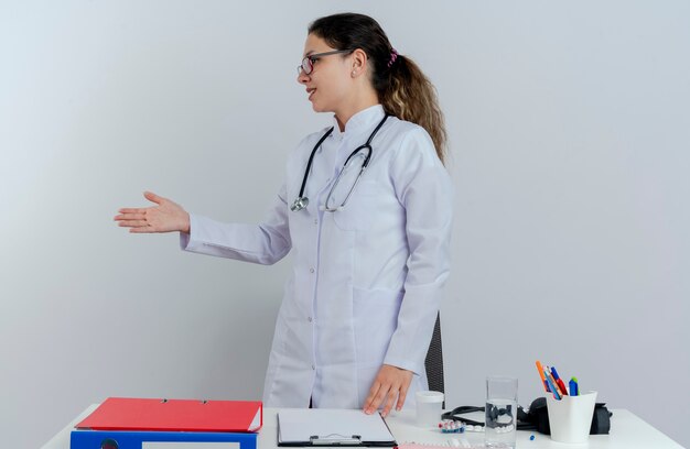 医療用ローブと聴診器と眼鏡を身に着けている印象的な若い女性医師が机の後ろに立って、医療ツールが頭を左右に回して挨拶ジェスチャーを分離
