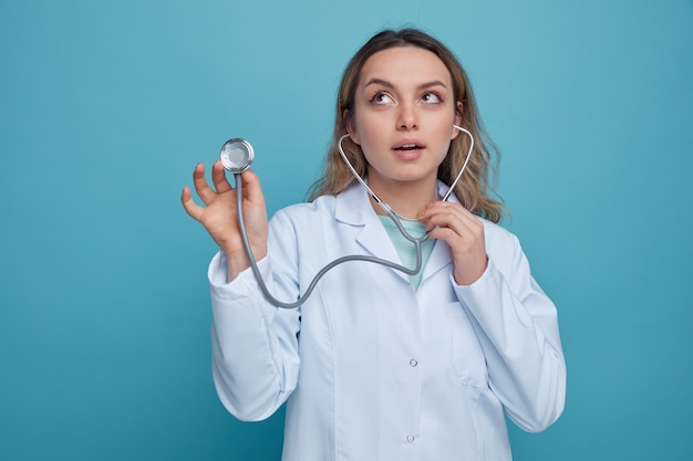 Impressed young female doctor wearing medical robe and stethoscope grabbing stethoscope looking up 