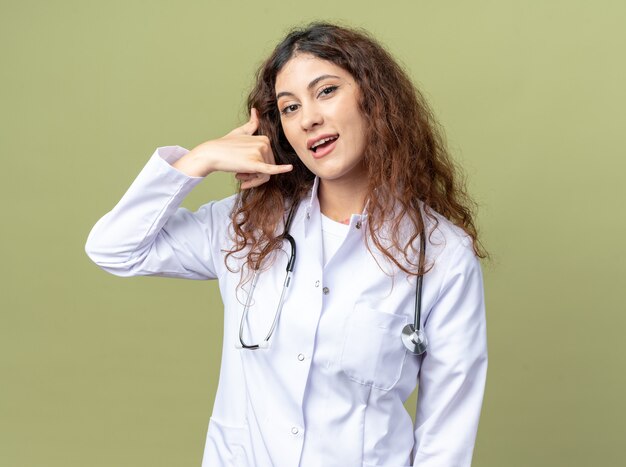 의료 가운과 청진기를 입고 올리브 녹색 벽에 격리된 통화 제스처를 하는 앞에서 보고 있는 젊은 여성 의사